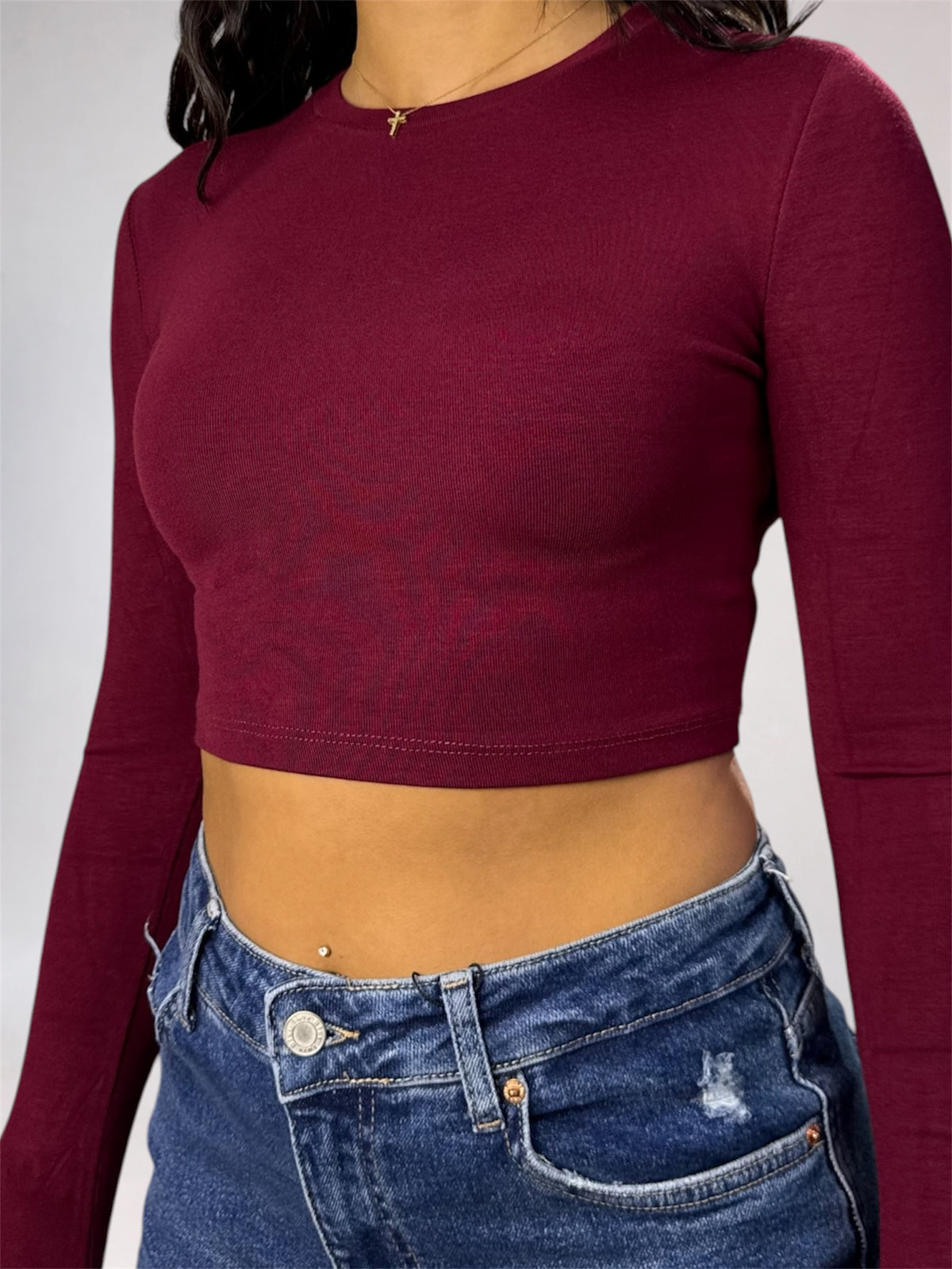 A woman wearing a fitted long-sleeve crop top in deep burgundy, paired with blue jeans, shown from the neck down against a light gray studio background.