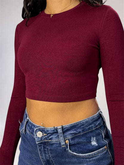 A woman wearing a fitted long-sleeve crop top in deep burgundy, paired with blue jeans, shown from the neck down against a light gray studio background.