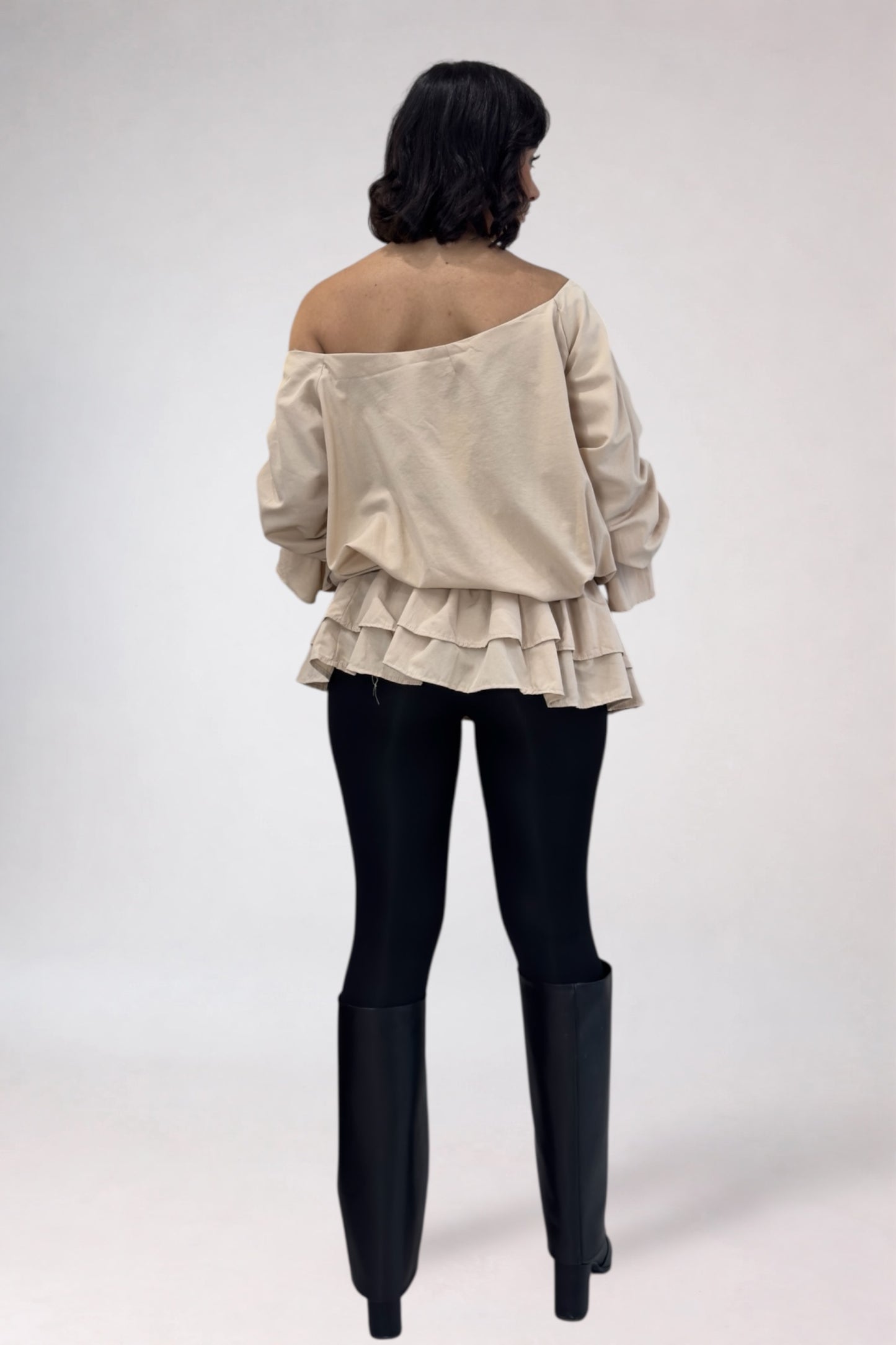 Woman wearing a beige off-shoulder top and black leggings with black boots on a white background