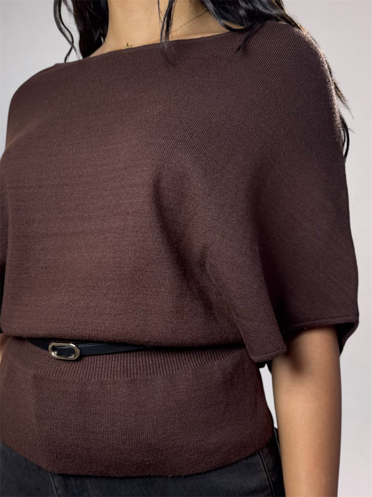 A close-up of a woman wearing a dark brown knit top with a wide boat neckline and short draped sleeves, styled with a thin black belt.