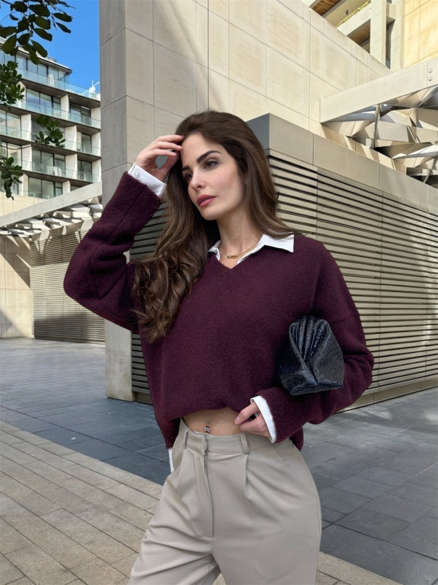 Woman in a burgundy sweater and beige pants standing outdoors near modern buildings.