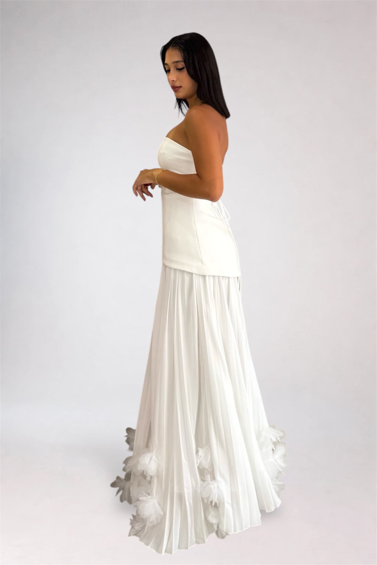 A woman modeling a strapless white maxi dress with a structured bodice and a flowing pleated skirt featuring feathered detailing at the hem, photographed against a plain light gray studio background.