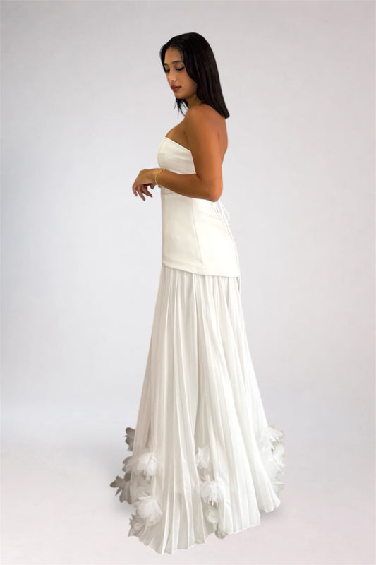 A woman modeling a strapless white maxi dress with a structured bodice and a flowing pleated skirt featuring feathered detailing at the hem, photographed against a plain light gray studio background.
