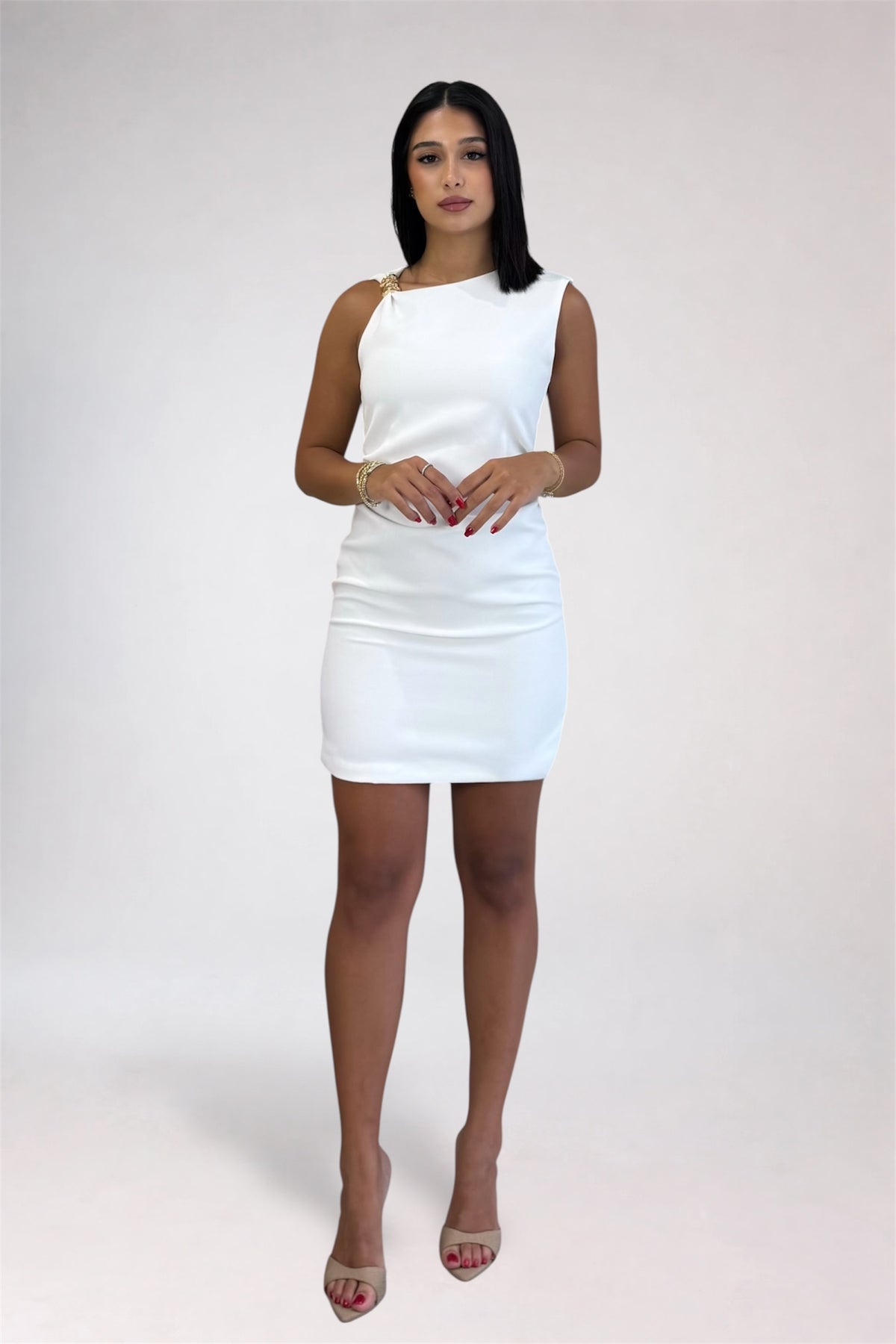 A woman modeling a white sleeveless bodycon mini dress with a single gold-tone ring detail at the shoulder, paired with nude high-heeled sandals, against a light gray studio background.