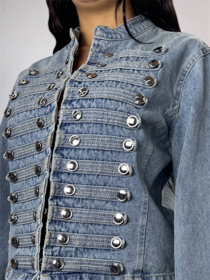 A close-up of a light blue denim jacket featuring multiple rows of silver metallic snap buttons and decorative stitching on the front, worn by a model.