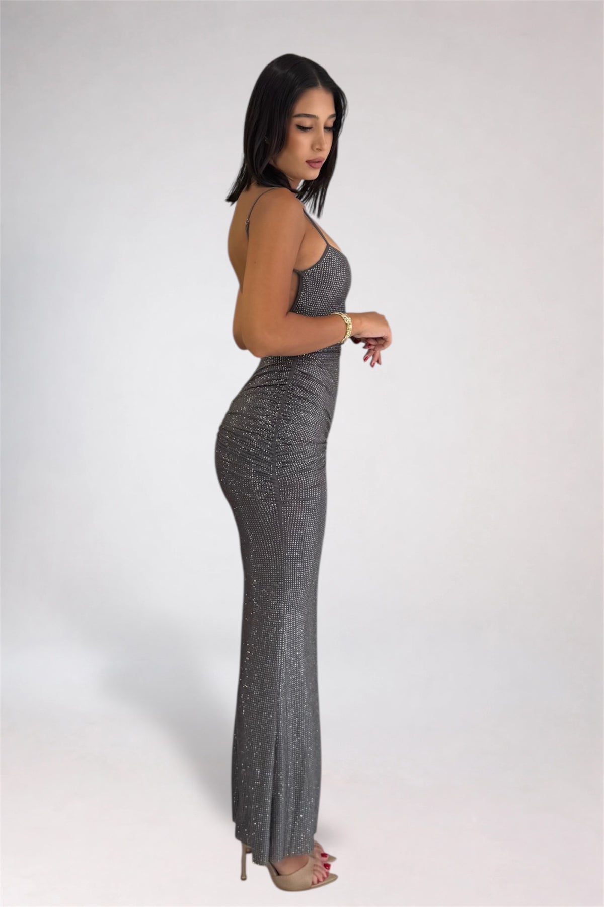 A woman modeling a sparkly, sleeveless, bodycon maxi dress in a shimmering gray color, standing in profile against a light gray studio background.