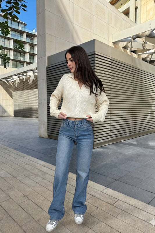 Woman in a cream sweater and blue jeans standing outdoors near modern architectural elements.