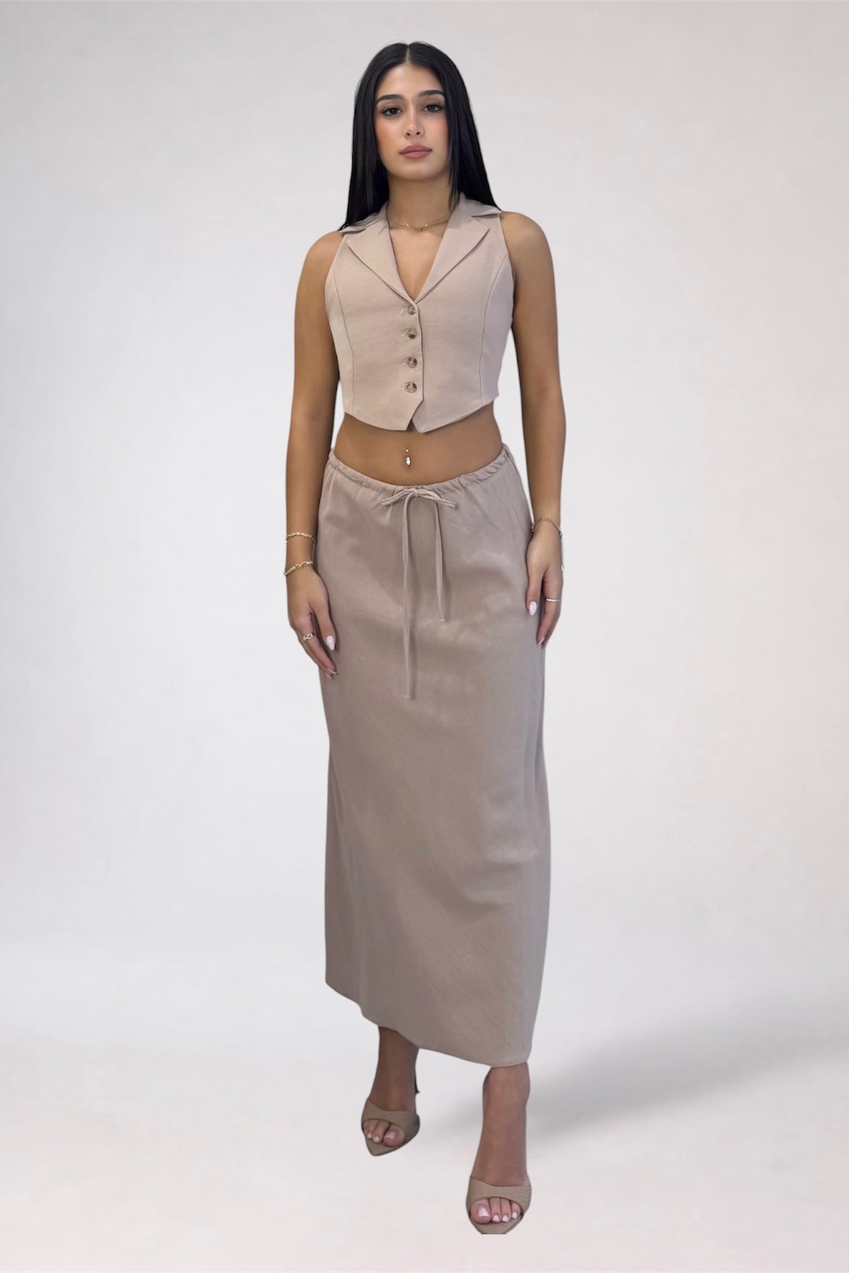 A model wearing a beige two-piece outfit consisting of a sleeveless collared crop top with buttons and a matching high-waisted drawstring midi skirt, set against a plain light gray studio background.