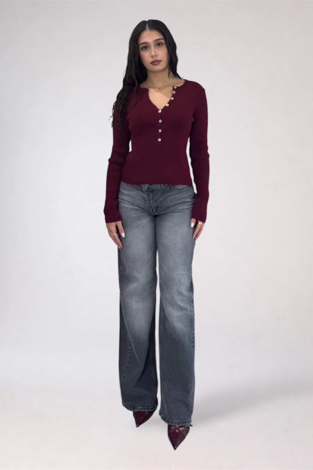 A woman modeling a burgundy long-sleeve knit top with decorative buttons down the front, paired with gray wide-leg jeans and dark pointed shoes, against a light gray studio background.