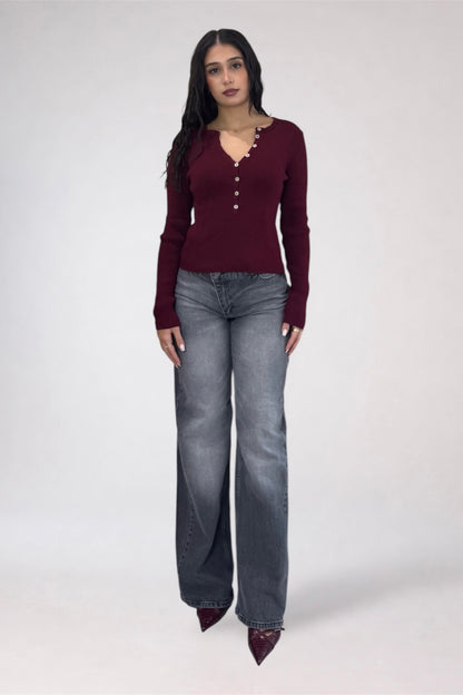 A woman modeling a burgundy long-sleeve knit top with decorative buttons down the front, paired with gray wide-leg jeans and dark pointed shoes, against a light gray studio background.