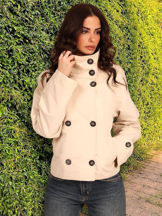 Woman wearing a beige coat with black buttons against a green hedge.