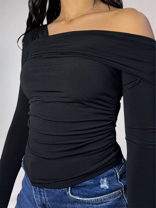 A close-up of a woman wearing a black off-the-shoulder long-sleeve top with ruched detailing, paired with blue jeans, against a light gray background.
