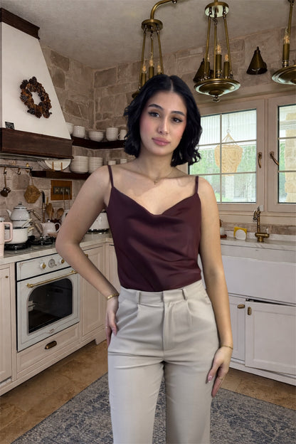 Woman standing in a kitchen wearing a brown top and beige pants.