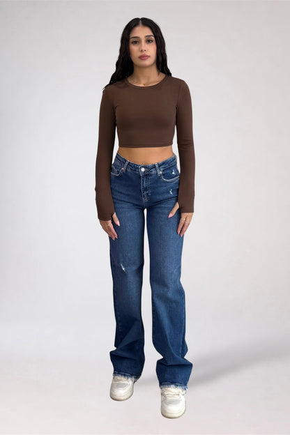 A woman modeling a brown long-sleeve crop top and blue high-waisted straight-leg jeans with white sneakers, standing against a light gray studio background.