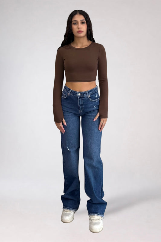 A woman modeling a brown long-sleeve crop top and blue high-waisted straight-leg jeans with white sneakers, standing against a light gray studio background.