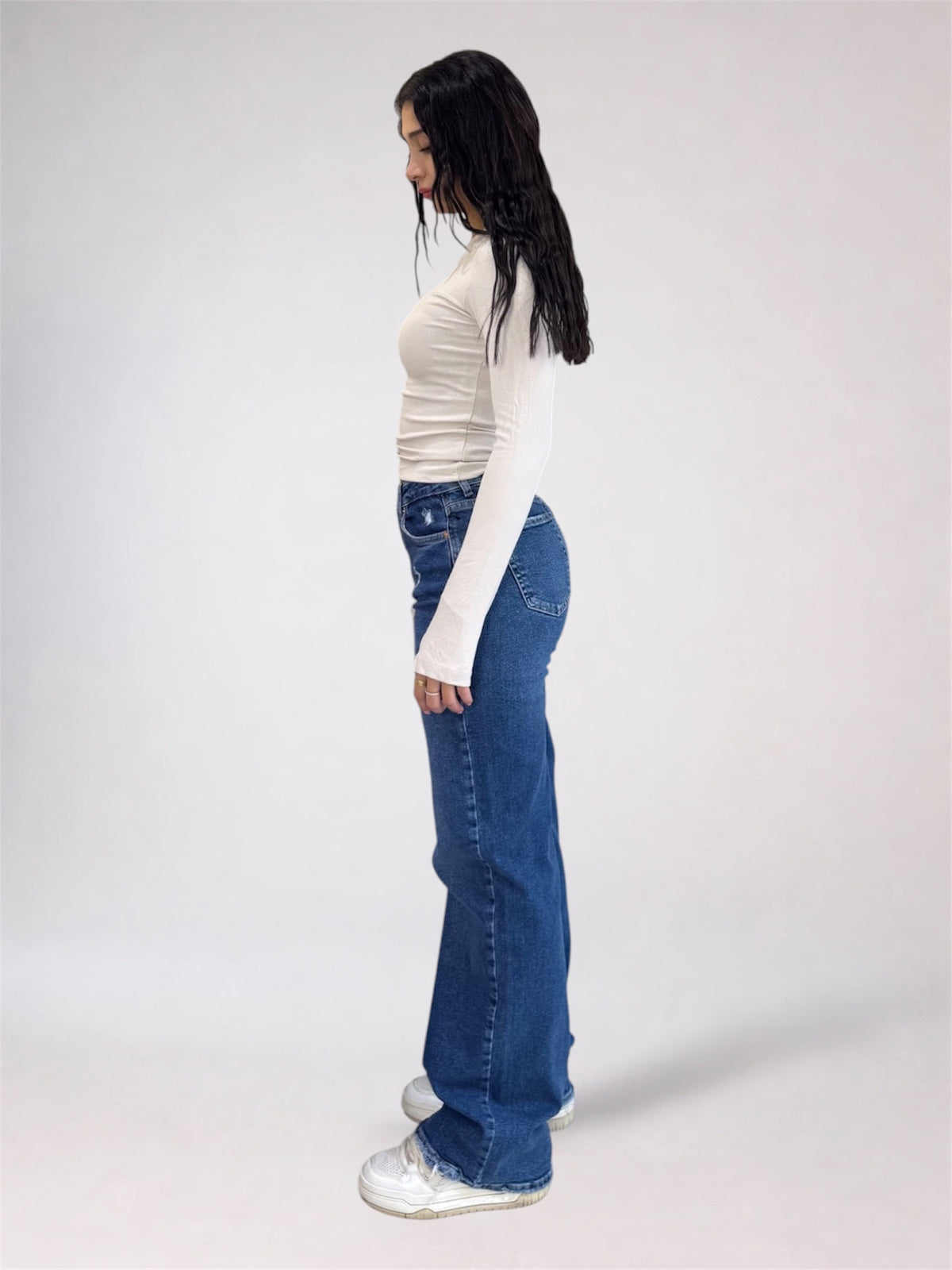 A woman shown in profile wearing a fitted white long-sleeve top, blue wide-leg jeans, and white sneakers against a plain light gray studio background.