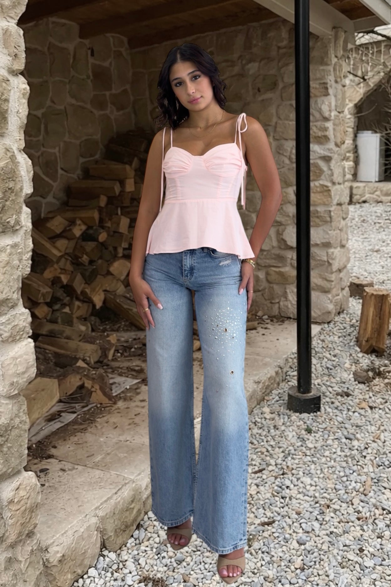Woman wearing a pink top and blue jeans standing in front of a stone building with wooden logs.