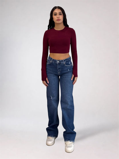A woman modeling a dark burgundy long-sleeve crop top and blue jeans with white sneakers, standing against a light gray studio background.