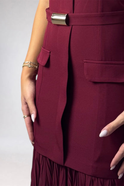 Close-up of a model wearing a sleeveless burgundy dress with a matching waist belt featuring a metallic gold buckle, side pockets, and a pleated skirt hem.