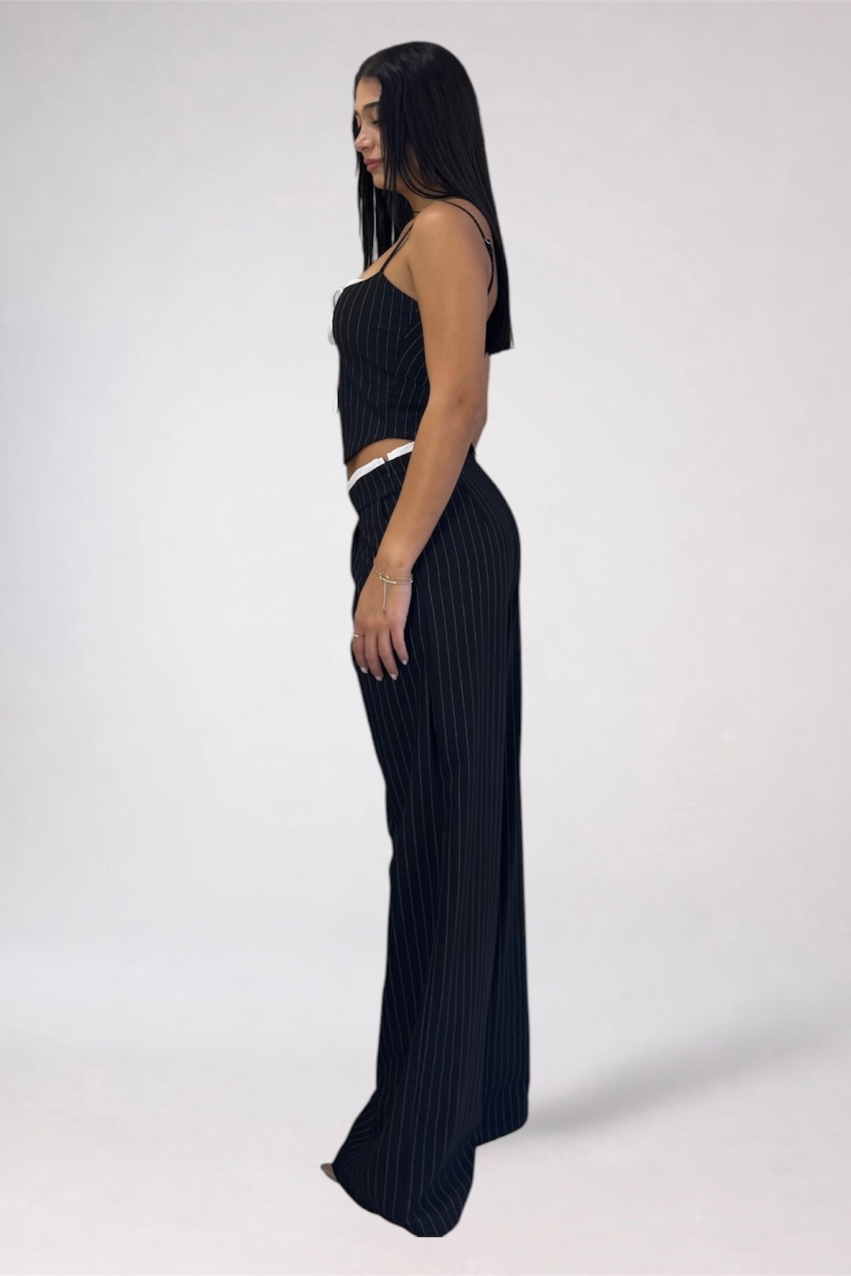 A woman stands in profile wearing a black ribbed knit two-piece outfit consisting of a halter crop top and matching wide-leg pants, photographed against a plain light gray studio background.