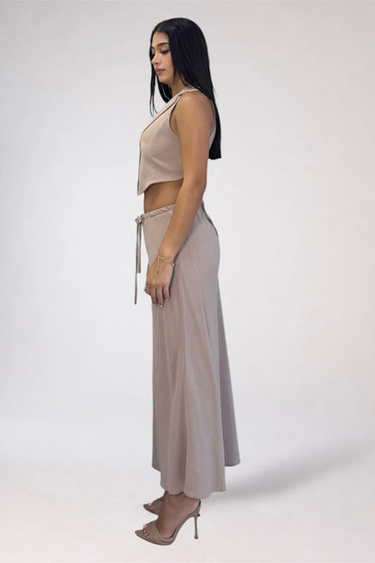 A woman stands in profile wearing a two-piece beige outfit consisting of a sleeveless crop top and a long skirt with a tie detail at the waist, paired with nude high heels, against a plain light gray studio background.
