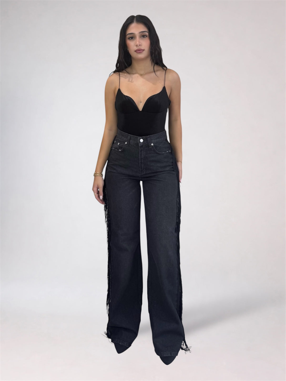 A woman modeling a black sweetheart-neckline camisole top and high-waisted, wide-leg black denim jeans with fringe detailing along the outer sides, set against a plain light gray studio background.