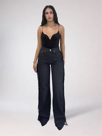 A woman modeling a black sweetheart-neckline camisole top and high-waisted, wide-leg black denim jeans with fringe detailing along the outer sides, set against a plain light gray studio background.