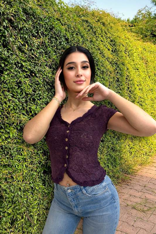 Woman in a purple top and blue jeans standing against a green hedge.