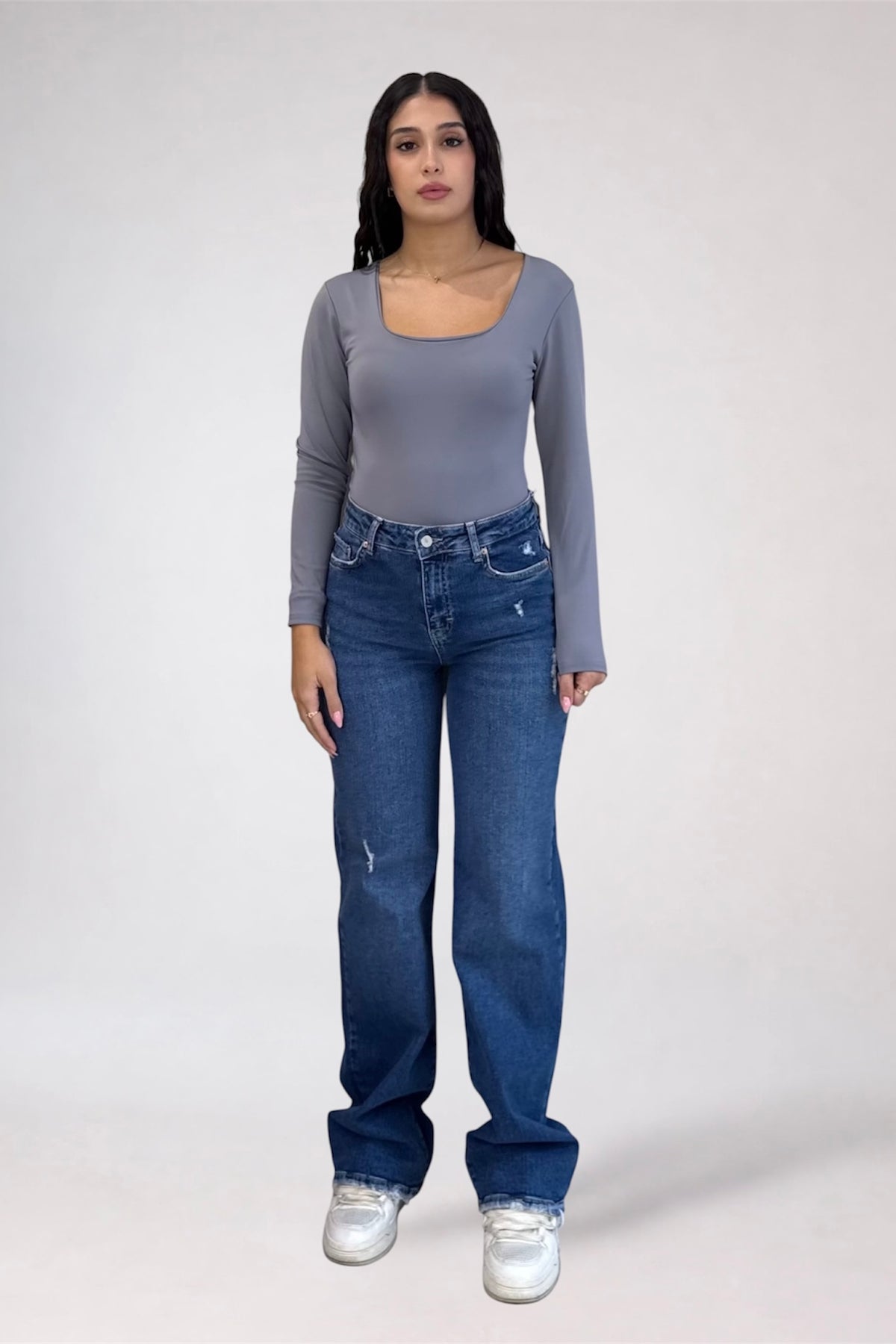 A woman modeling a gray long-sleeve scoop-neck top and blue jeans against a plain light gray studio background.
