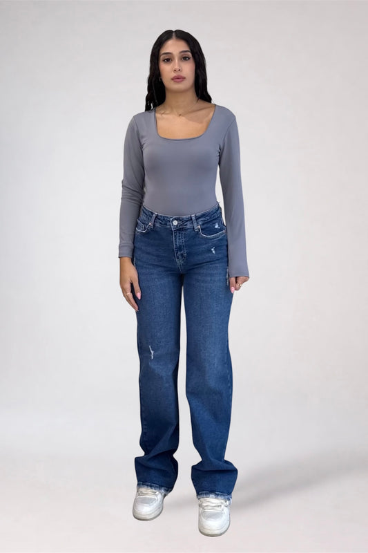A woman modeling a gray long-sleeve scoop-neck top and blue jeans against a plain light gray studio background.