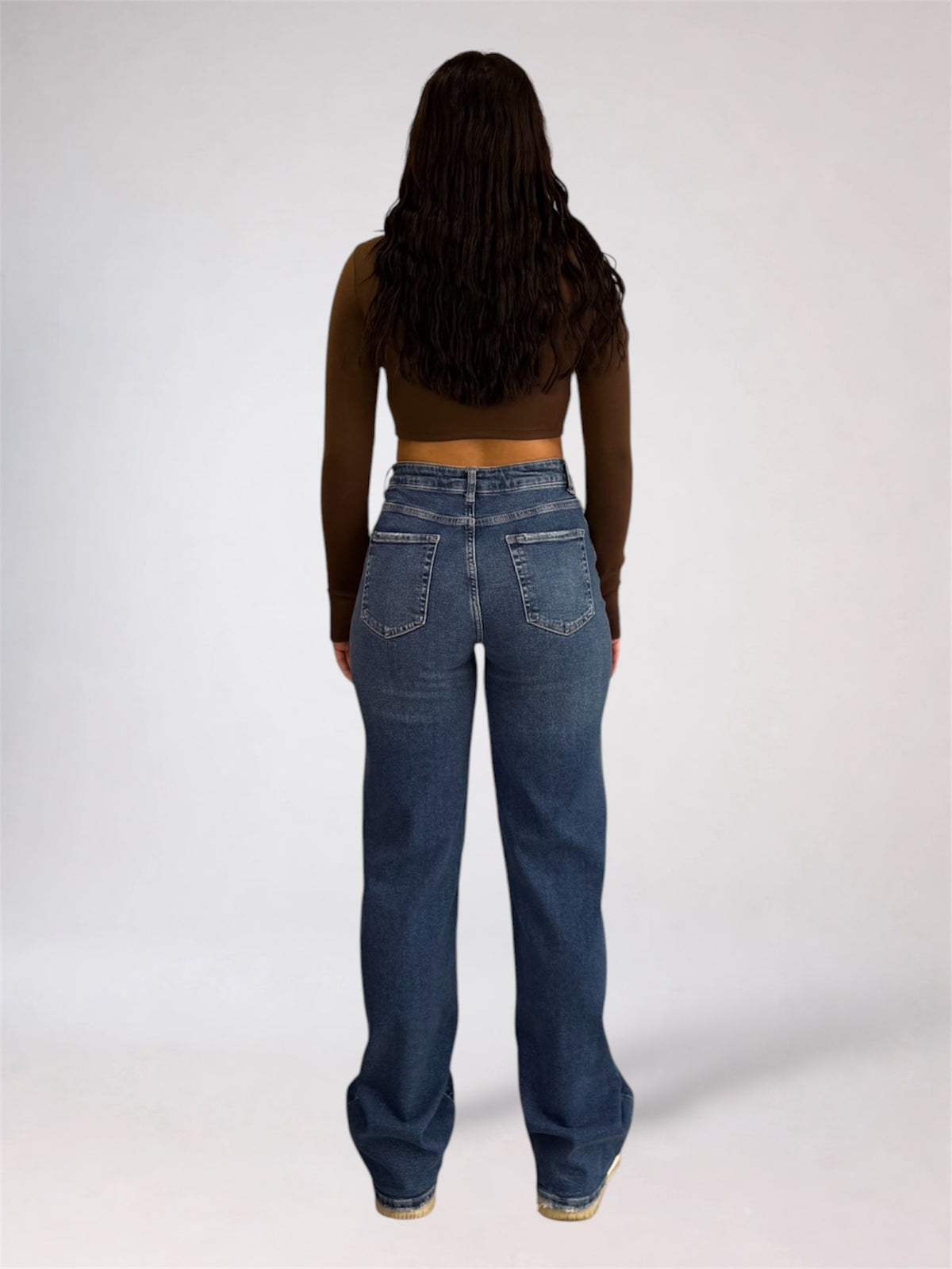 A woman viewed from behind wearing a dark brown long-sleeve crop top and high-waisted, wide-leg blue denim jeans against a plain light gray studio background.