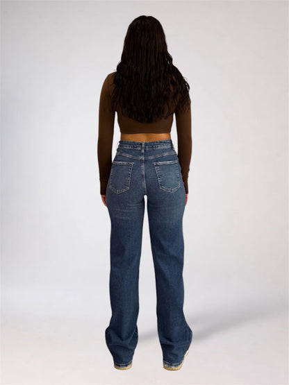 A woman viewed from behind wearing a dark brown long-sleeve crop top and high-waisted, wide-leg blue denim jeans against a plain light gray studio background.