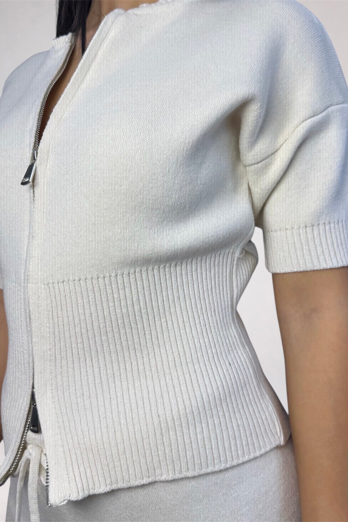 Close-up of a person wearing a short-sleeve knit zip-front top in a light off-white or cream color, with ribbed trim and a visible zipper detail.