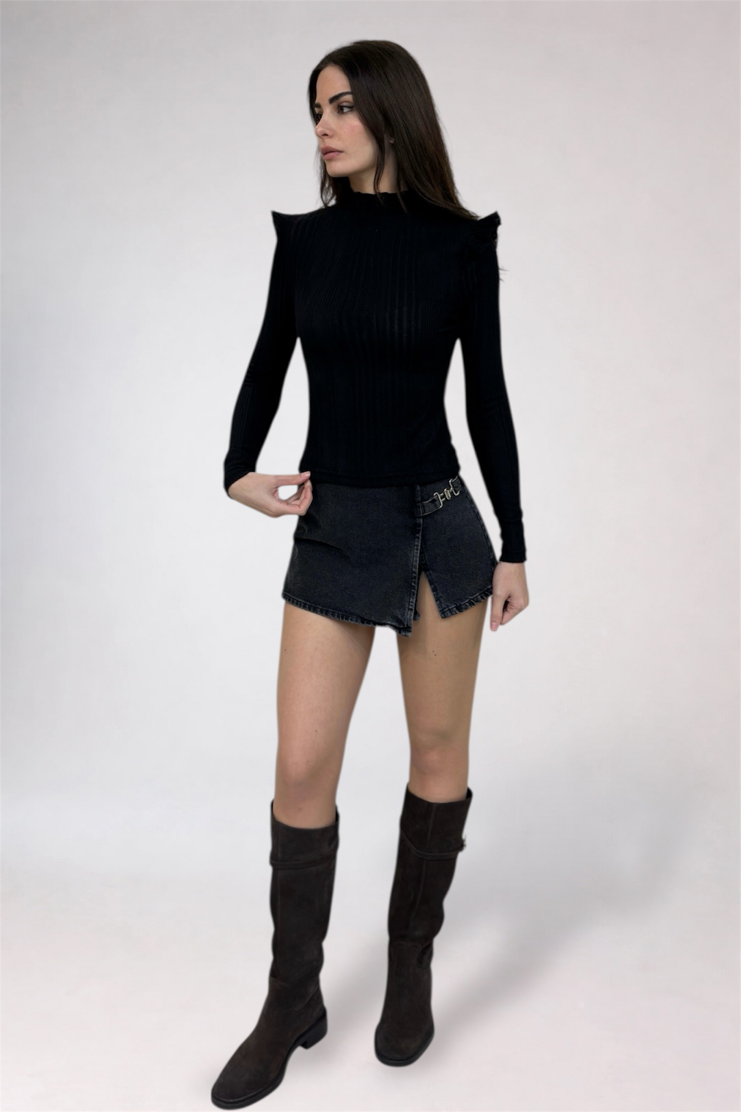 Woman wearing a black long-sleeve top and denim skirt with knee-high black boots on a white background
