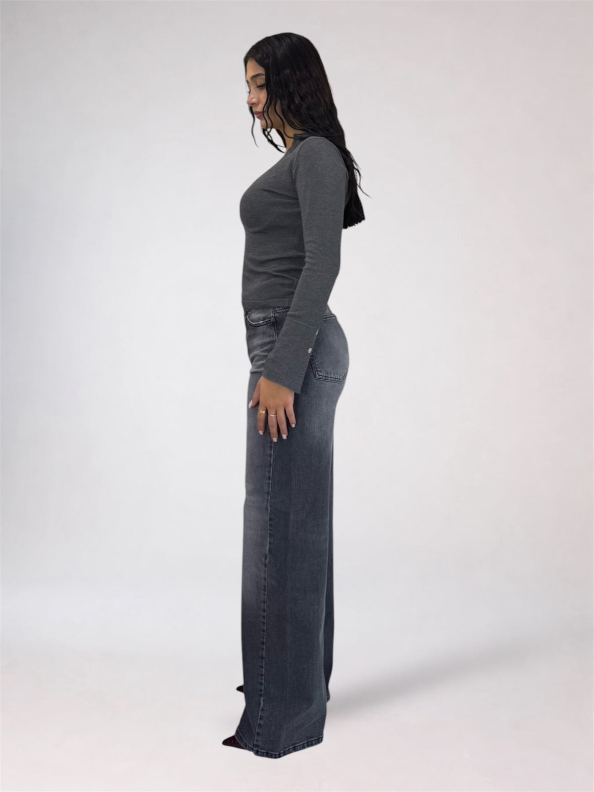 A woman shown in profile wearing a dark gray long-sleeve top and matching dark gray wide-leg jeans against a plain light gray studio background.