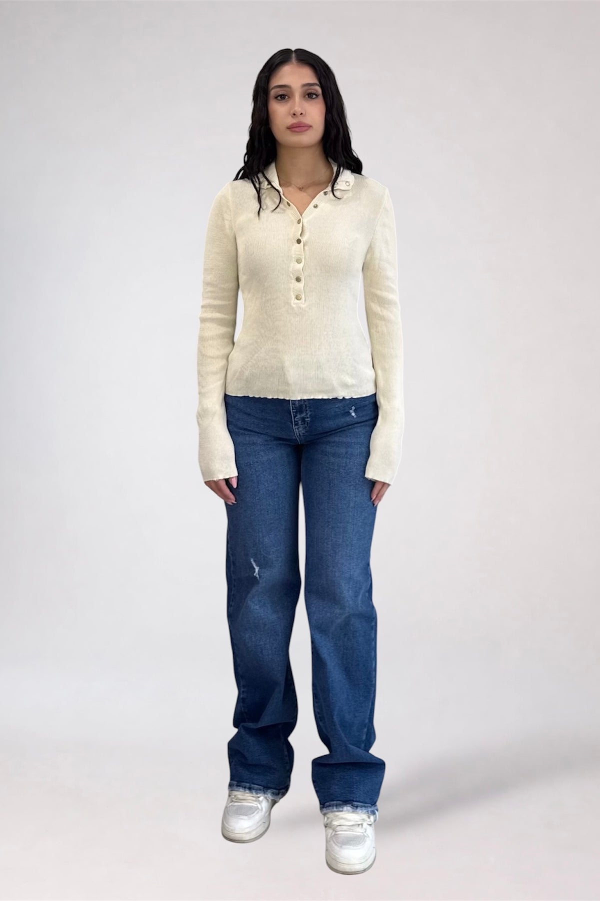 A woman stands against a light gray studio background wearing a cream ribbed knit long-sleeve henley top, blue jeans, and white sneakers.