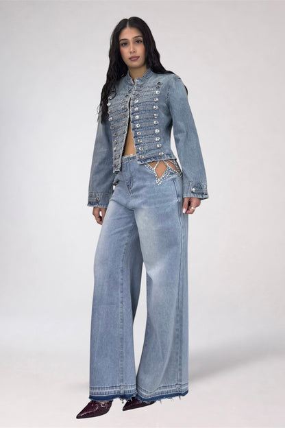 A model wearing a light blue denim set consisting of a cropped, embellished jacket with silver studs and matching wide-leg jeans, paired with dark pointed shoes, against a plain light gray studio background.