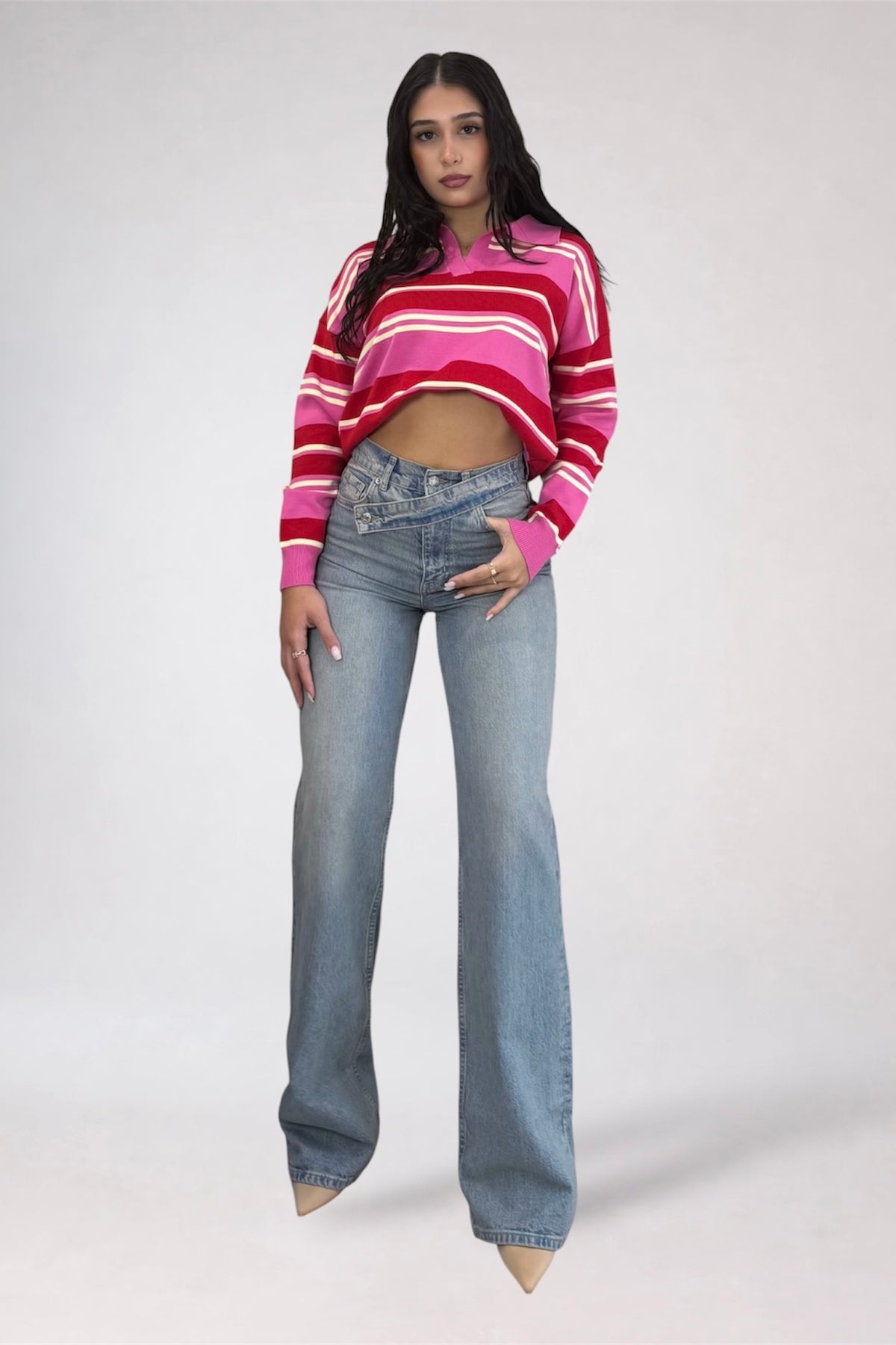 A woman modeling a pink and red striped cropped long-sleeve knit sweater paired with light-wash blue flare jeans, standing against a light gray studio background.