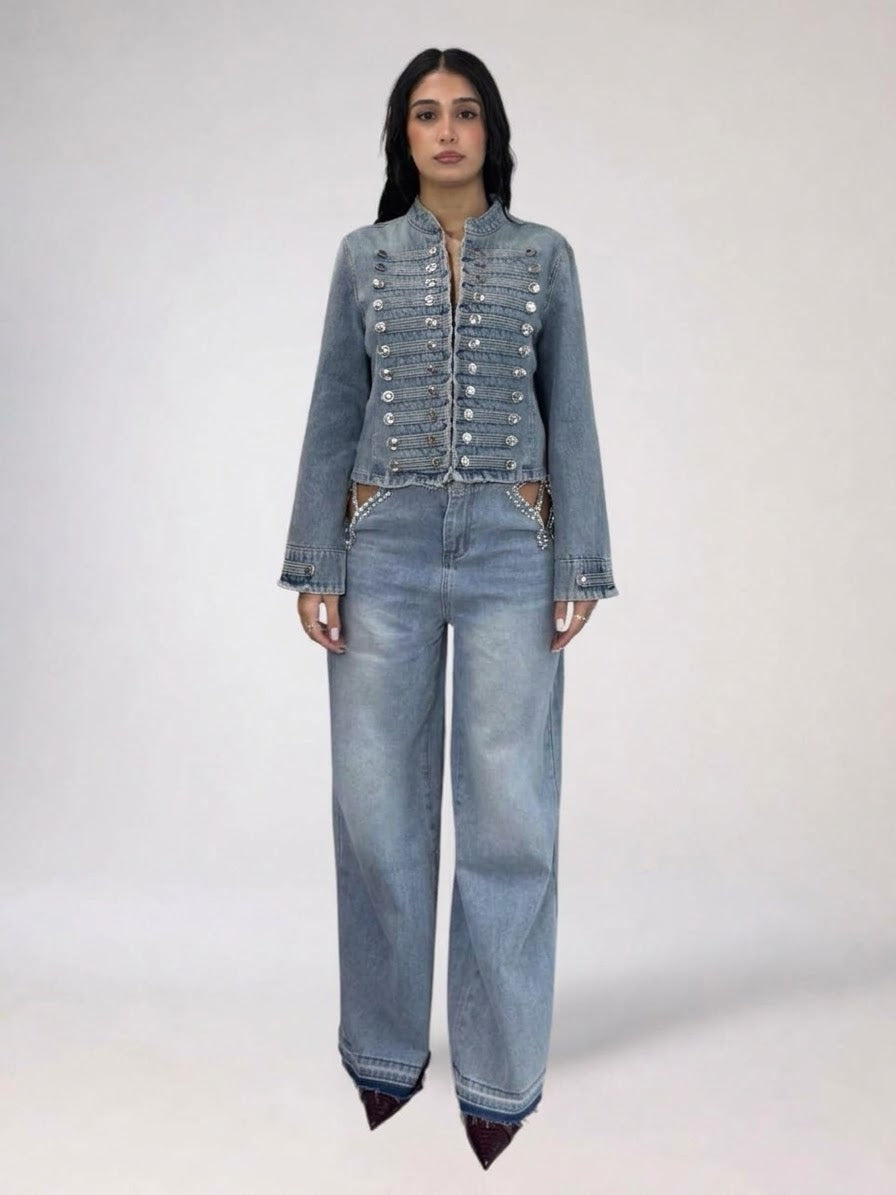 A model stands against a light gray studio background wearing a matching light-wash denim set consisting of a cropped, embellished jacket with silver button details and wide-leg jeans.
