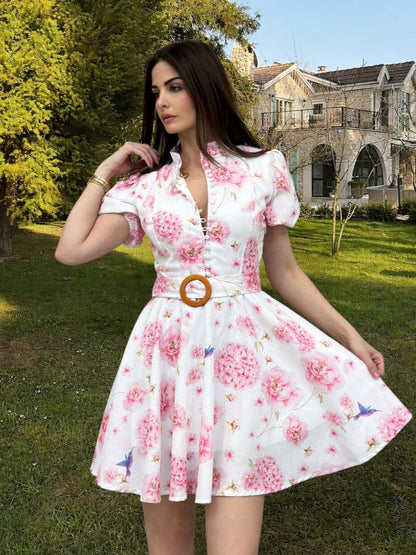 Woman wearing a floral dress in an outdoor setting with greenery and a building in the background