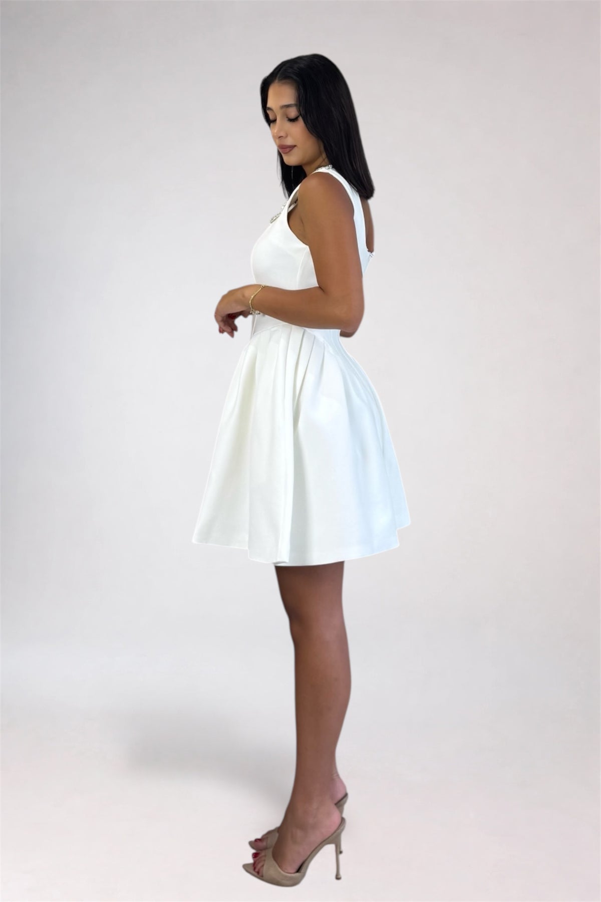 A woman modeling a short white sleeveless dress with a flared skirt, paired with nude high-heeled sandals, photographed against a plain light gray studio background.