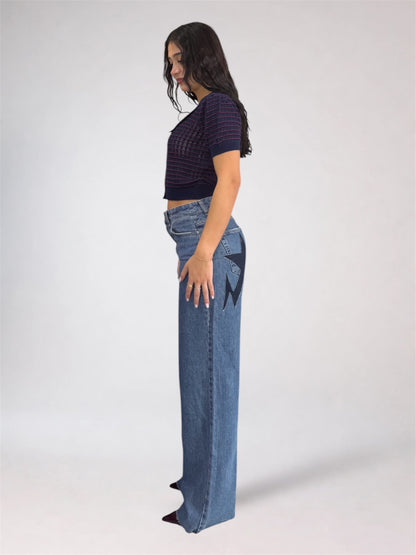A woman stands in profile wearing a dark purple short-sleeve knit top and high-waisted, wide-leg blue jeans with a black geometric pattern on the outer thigh, photographed against a light gray studio background.