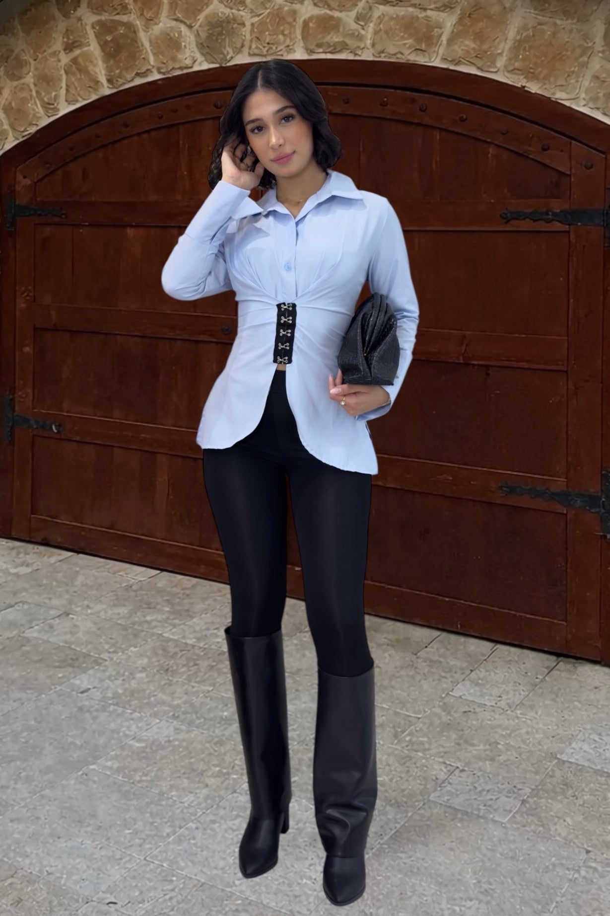 Woman in a white blouse with a black belt and black leggings standing in front of a stone wall.