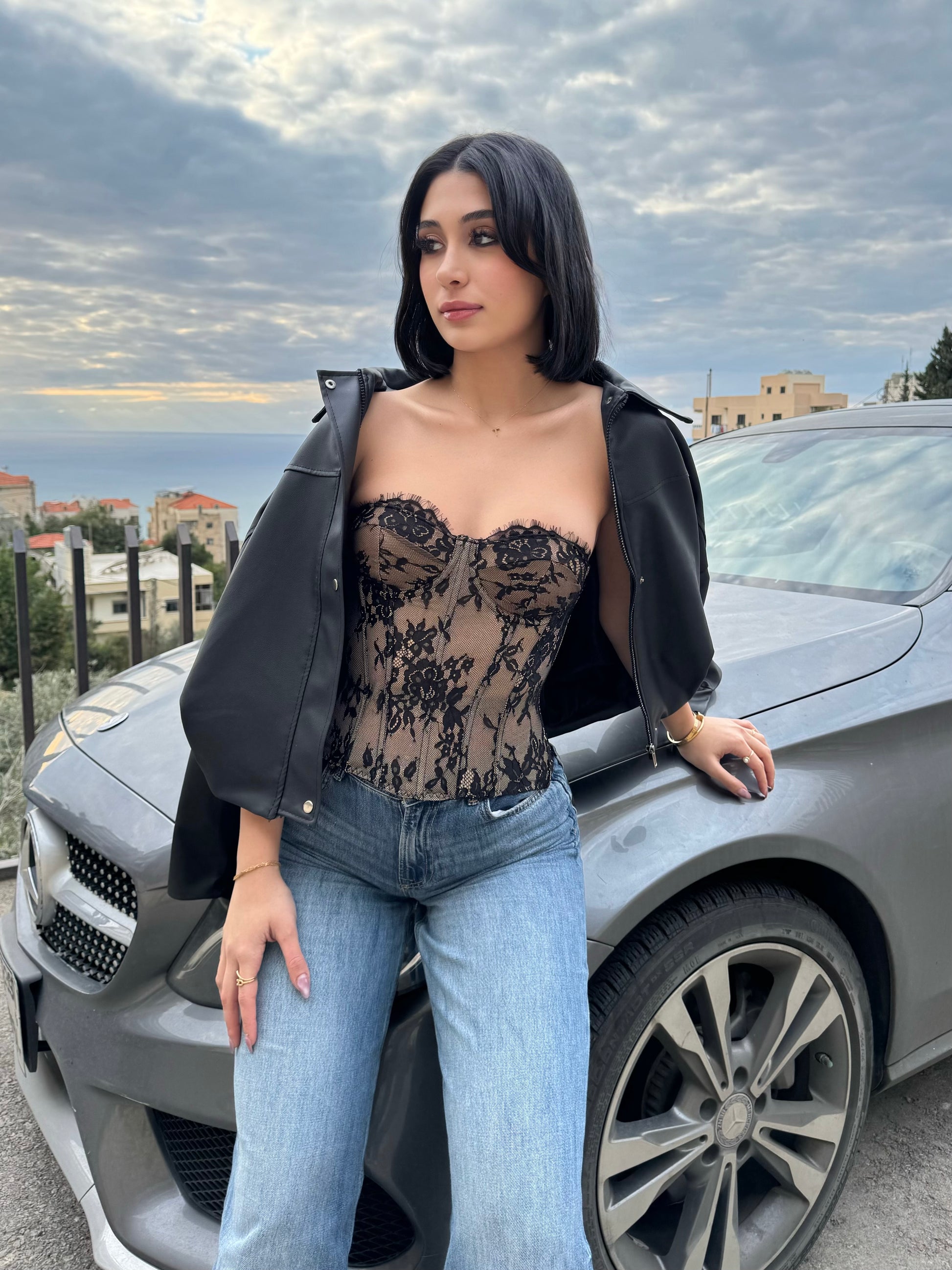 Woman standing next to a car wearing a black lace top and blue jeans with a cityscape in the background.