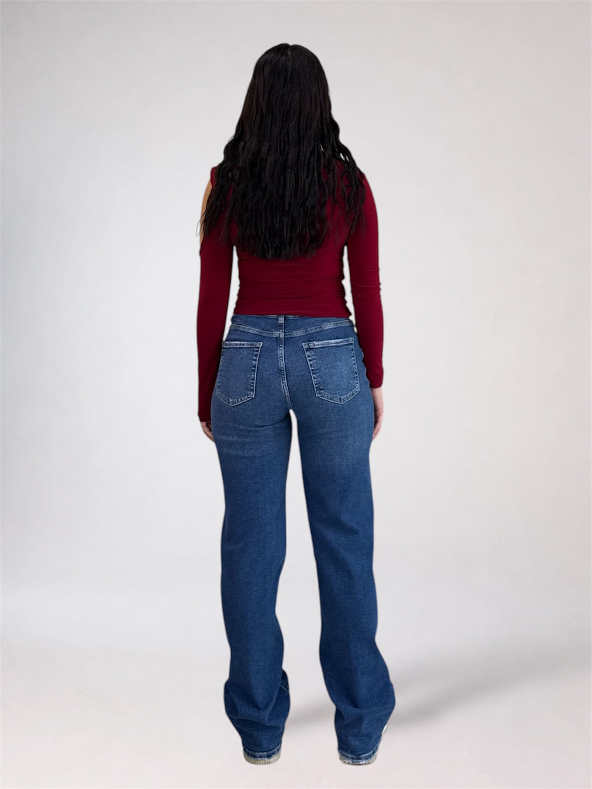 A woman viewed from behind wearing a dark red long-sleeve top and blue jeans, standing against a plain light gray studio background.