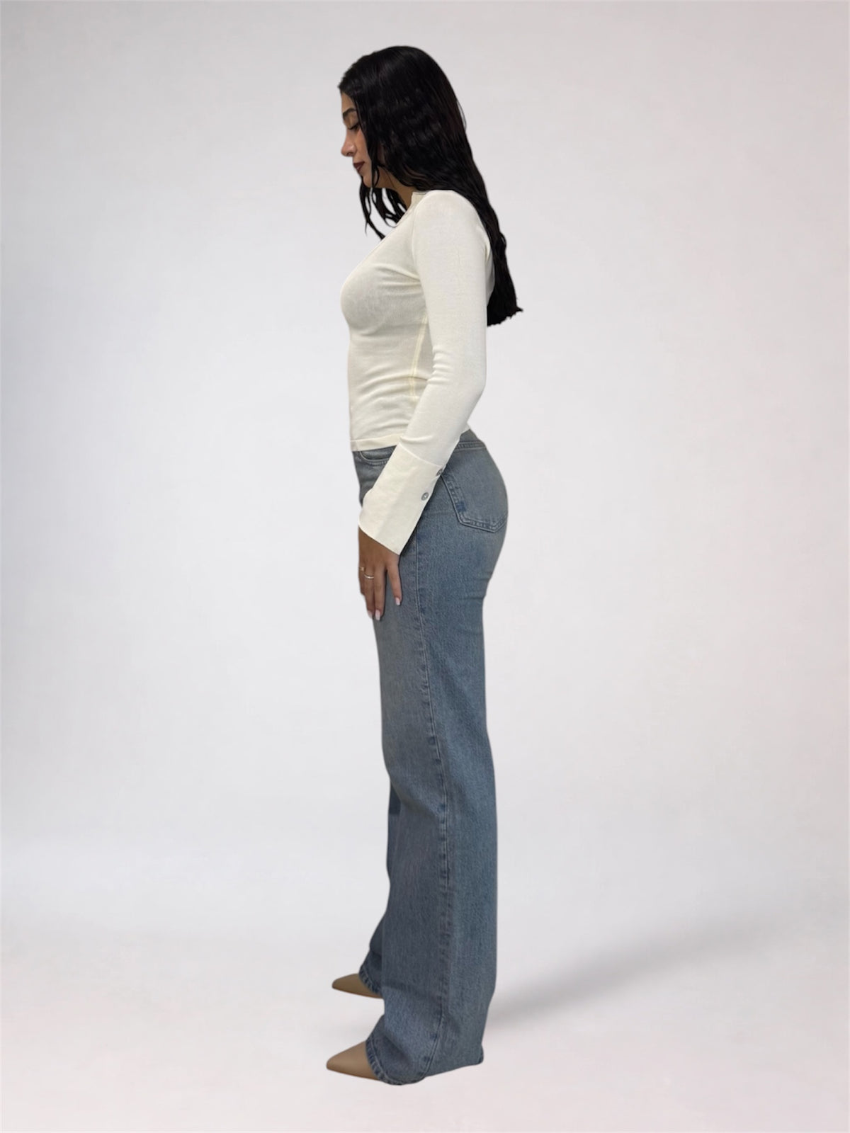A woman shown in profile wearing a fitted white long-sleeve top and light blue flared jeans against a plain light gray studio background.
