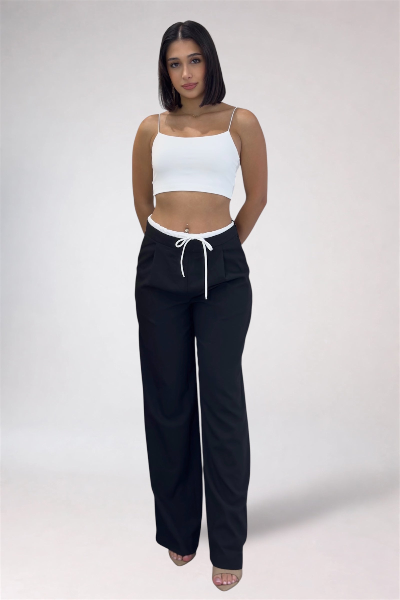 Woman wearing a white crop top and black pants with a drawstring on a plain background