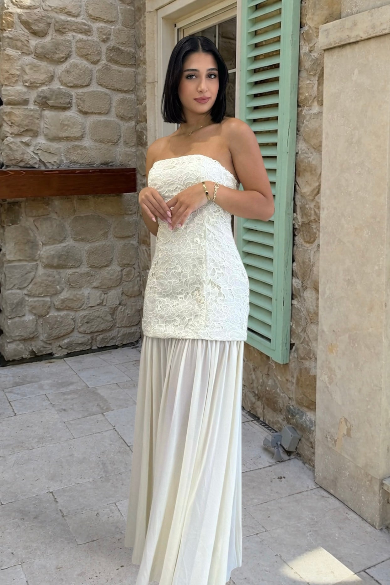 Woman in a white strapless dress standing in front of a stone wall with green shutters.