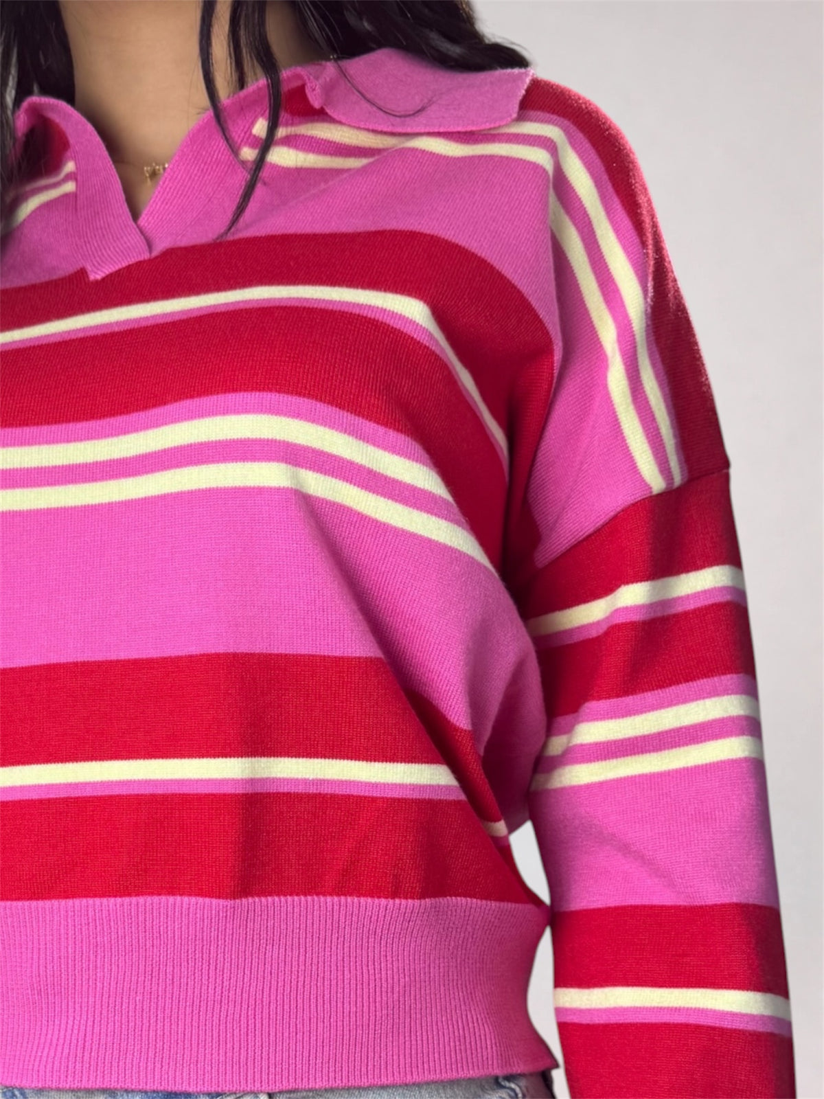 A close-up of a person wearing a knit long-sleeve polo-style top with bold horizontal stripes in pink, red, and cream, against a plain light gray background.