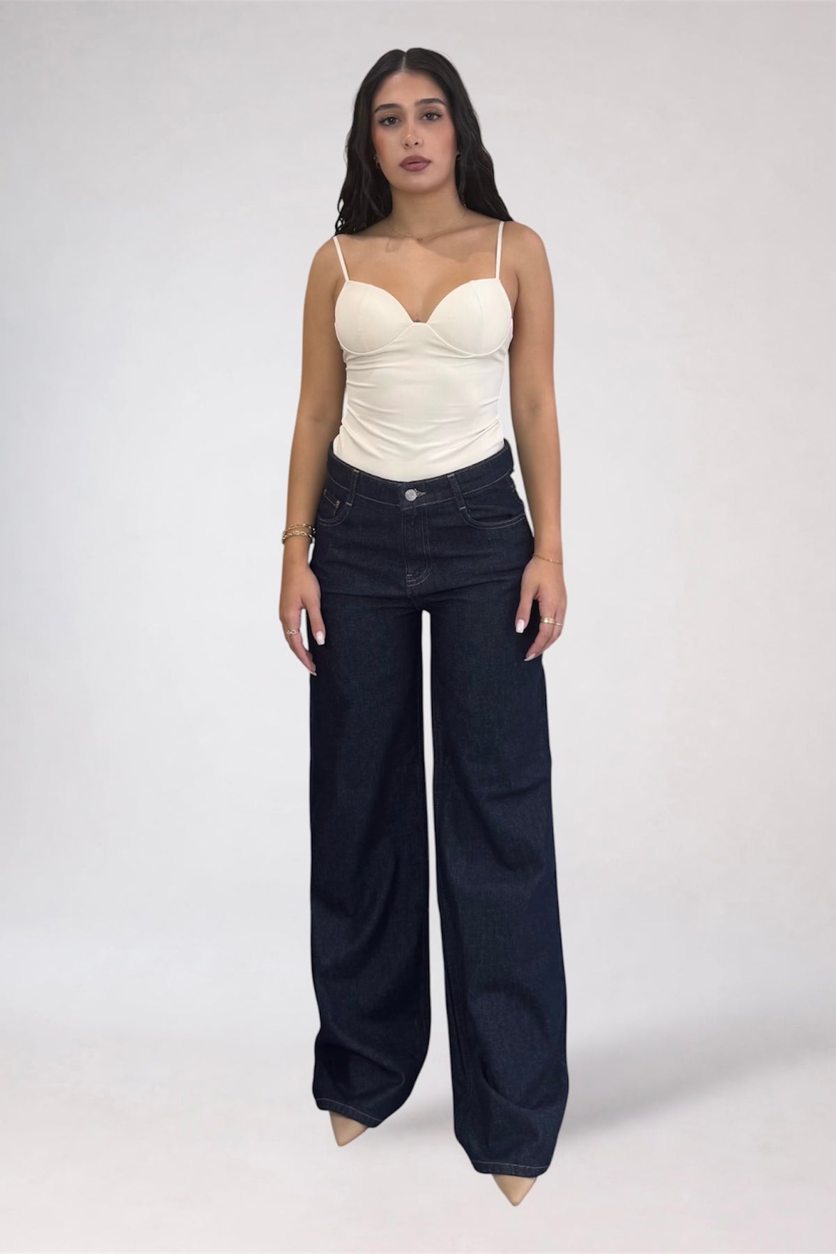 A woman modeling a cream-colored ribbed camisole top with spaghetti straps and dark wide-leg denim jeans against a plain light gray studio background.